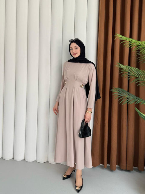 Woman in a beige dress and black hijab standing in front of white and brown curtains.