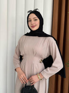 Woman wearing a beige dress and black hijab with sunglasses on her head, standing against a white curtain.