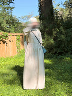 A person standing outdoors in a grassy yard wearing a long cream dress and a head covering, with a small blue shoulder bag. Trees, a wooden fence, and blue sky are visible in the background.