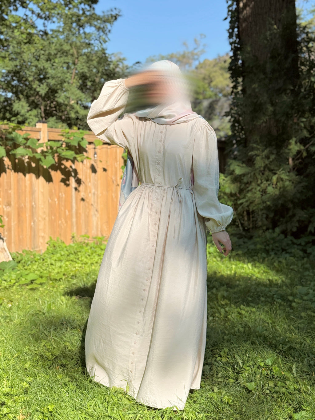 A person wearing a long cream-colored linen dress with a matching head covering, standing outdoors on a grassy lawn with a wooden fence and trees in the background.