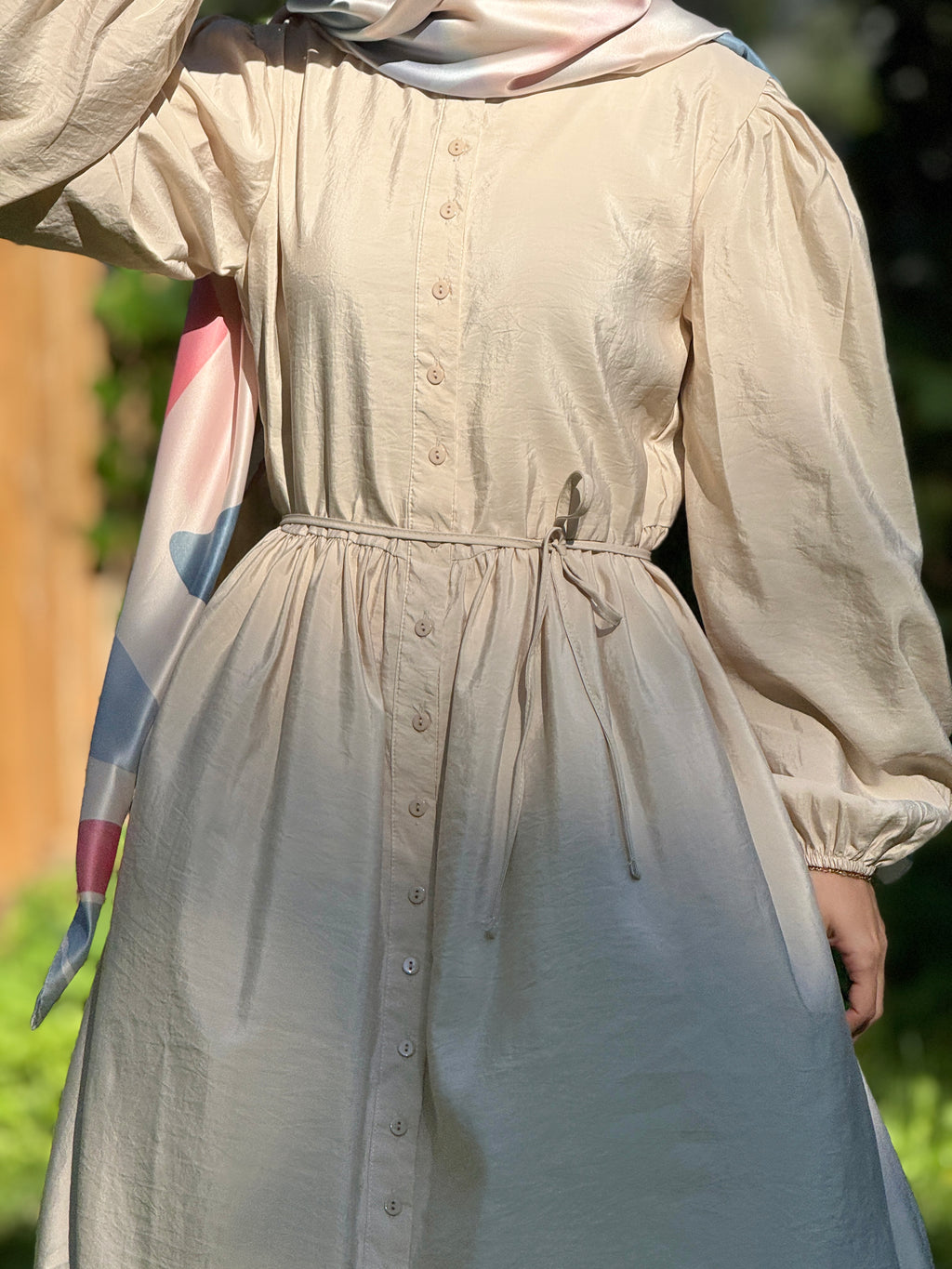 A person wearing a light beige long-sleeve dress with a button front, gathered waist, and drawstring tie, photographed outdoors with a soft-focus green and brown background.