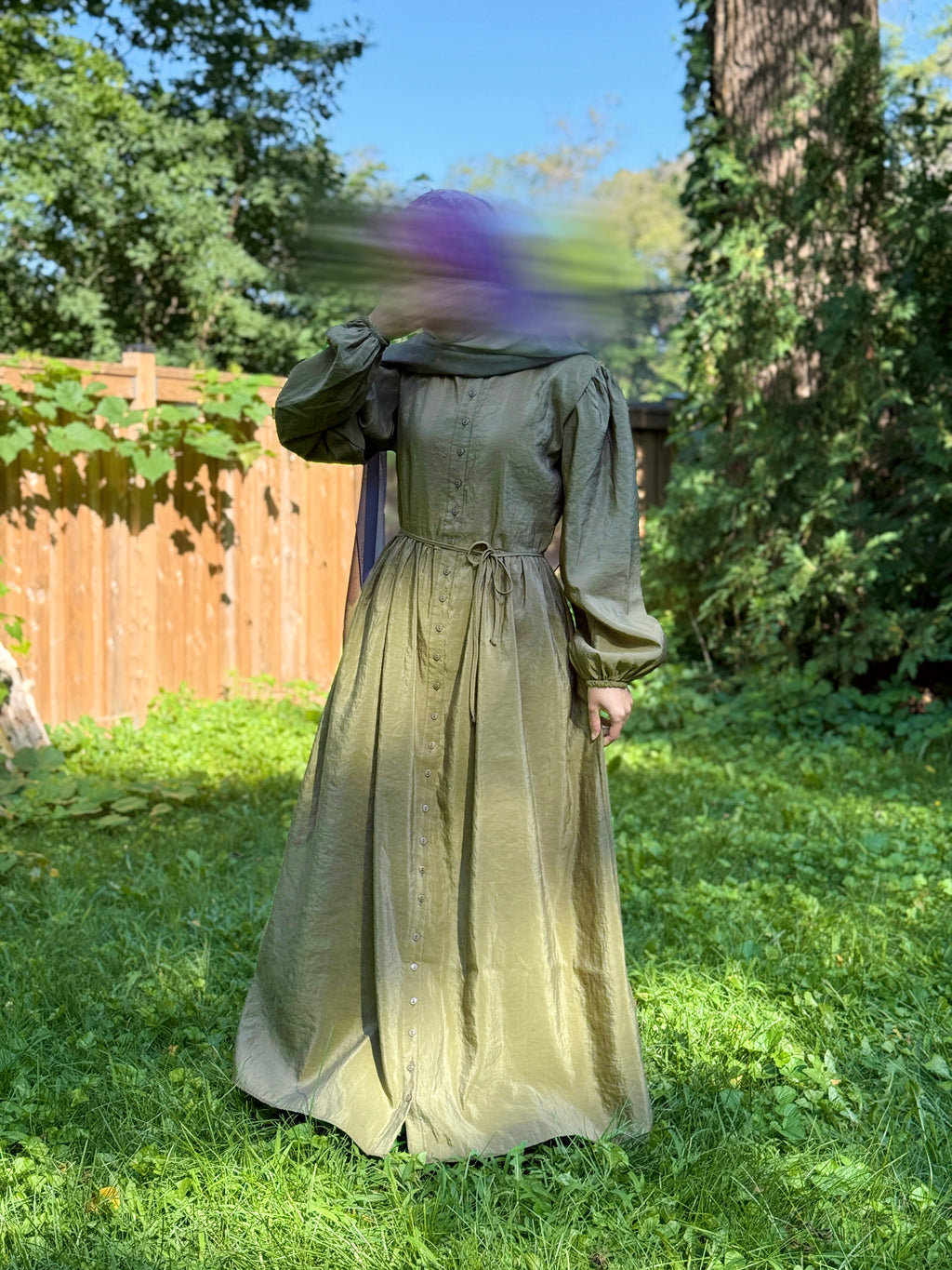 A person wearing a long-sleeved olive dress and head covering stands outdoors in a grassy backyard with a wooden fence and trees. The face is blurred, and there is motion blur across the upper body.