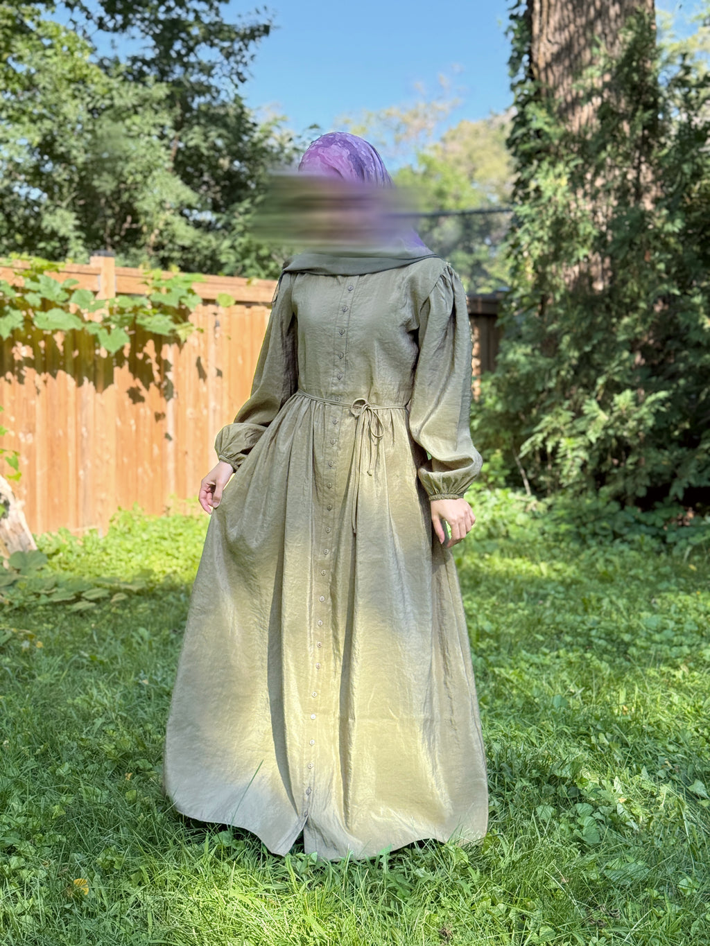 A person wearing a long-sleeved, floor-length dress in a muted olive green color, standing in a grassy backyard with a wooden fence and trees in the background.