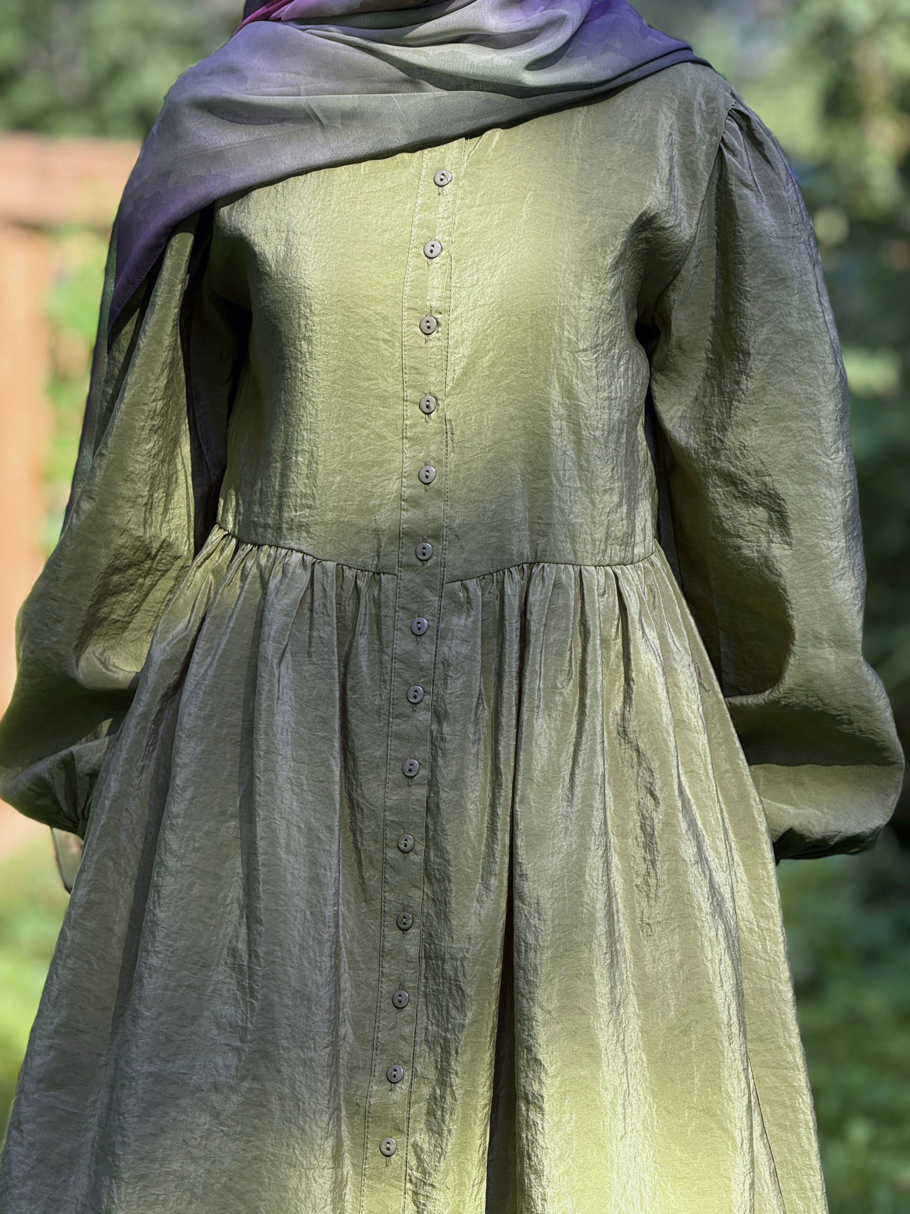 A closeup of long-sleeved dress in olive green with a button-down front and a gathered waist, styled with a purple and green gradient scarf, shown outdoors against a blurred green background.