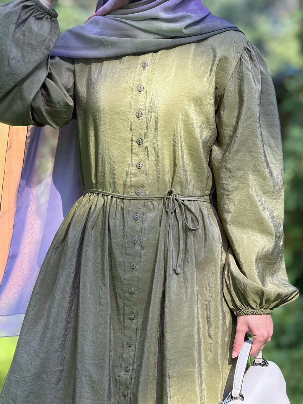 A person wearing a long-sleeved olive green dress with a button-down front and a matching drawstring waist, paired with a purple headscarf and holding a white handbag.