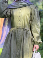 A person wearing a long-sleeved olive green dress with a button-down front and a matching drawstring waist, paired with a purple headscarf and holding a white handbag.