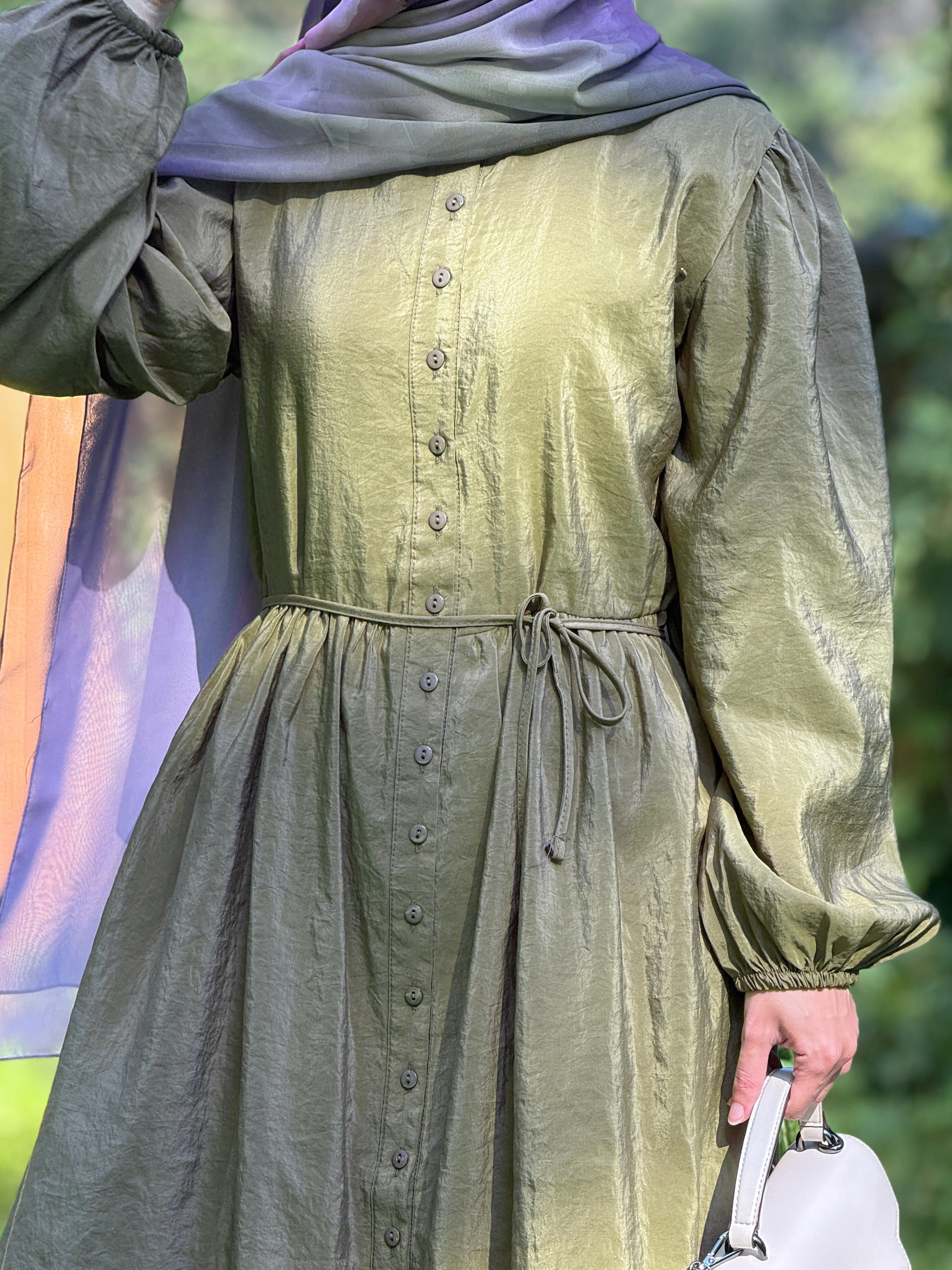 A person wearing a long-sleeved olive green dress with a button-down front and a matching drawstring waist, paired with a purple headscarf and holding a white handbag.