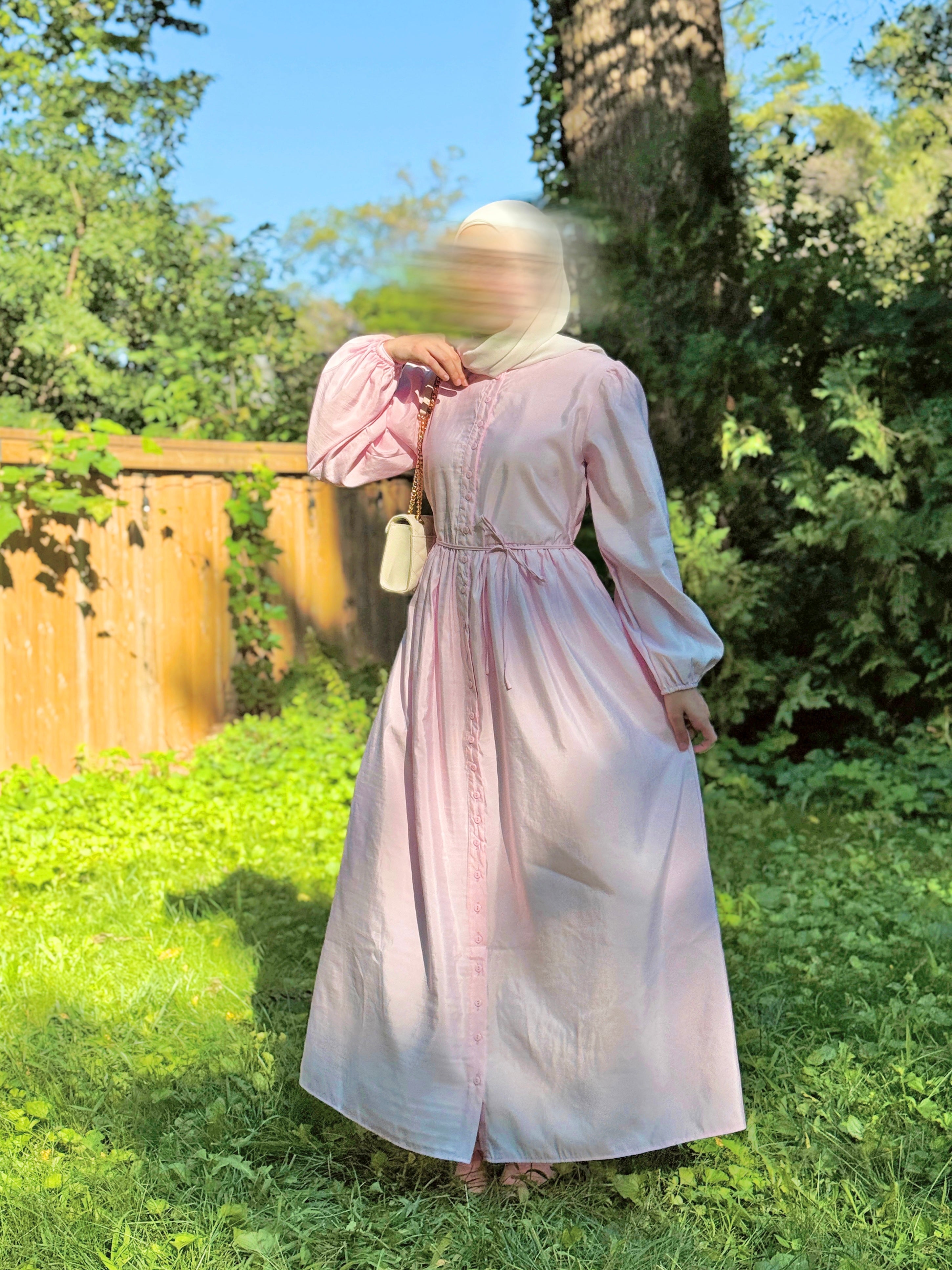 A woman wearing a long, light pink dress with long sleeves and a gathered waist, standing outdoors in a grassy yard with trees and a wooden fence in the background.
