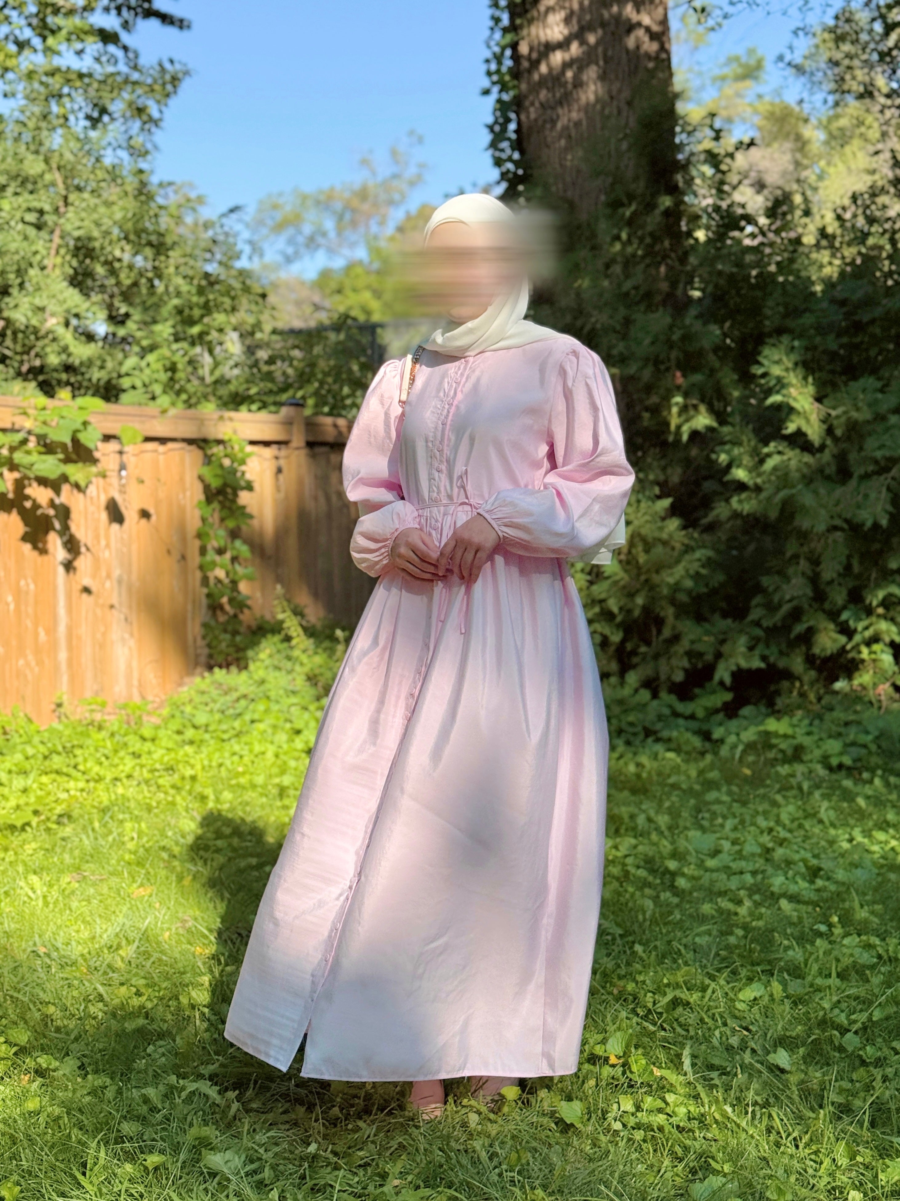 A person wearing a long light pink dress with a matching head covering stands outdoors on grass in a backyard with trees and a wooden fence visible.