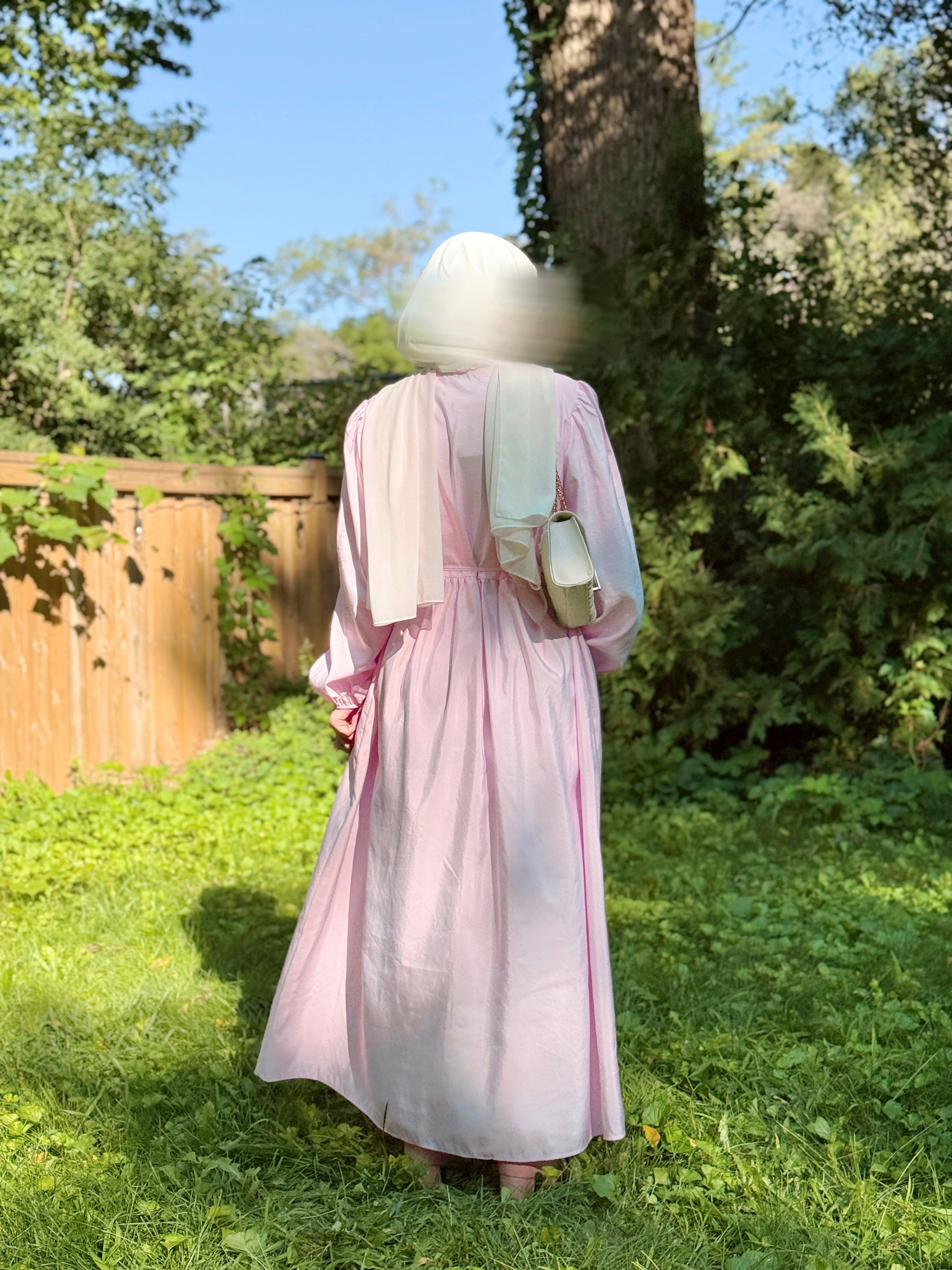 A person standing outdoors in a grassy yard wearing a long light pink dress with a white head covering and a small beige shoulder bag, photographed from the back.