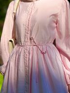 A close-up of a person wearing a light pink long-sleeve dress with a button front, gathered waist, and drawstring tie. A cream shoulder bag with a gold chain is partially visible.
