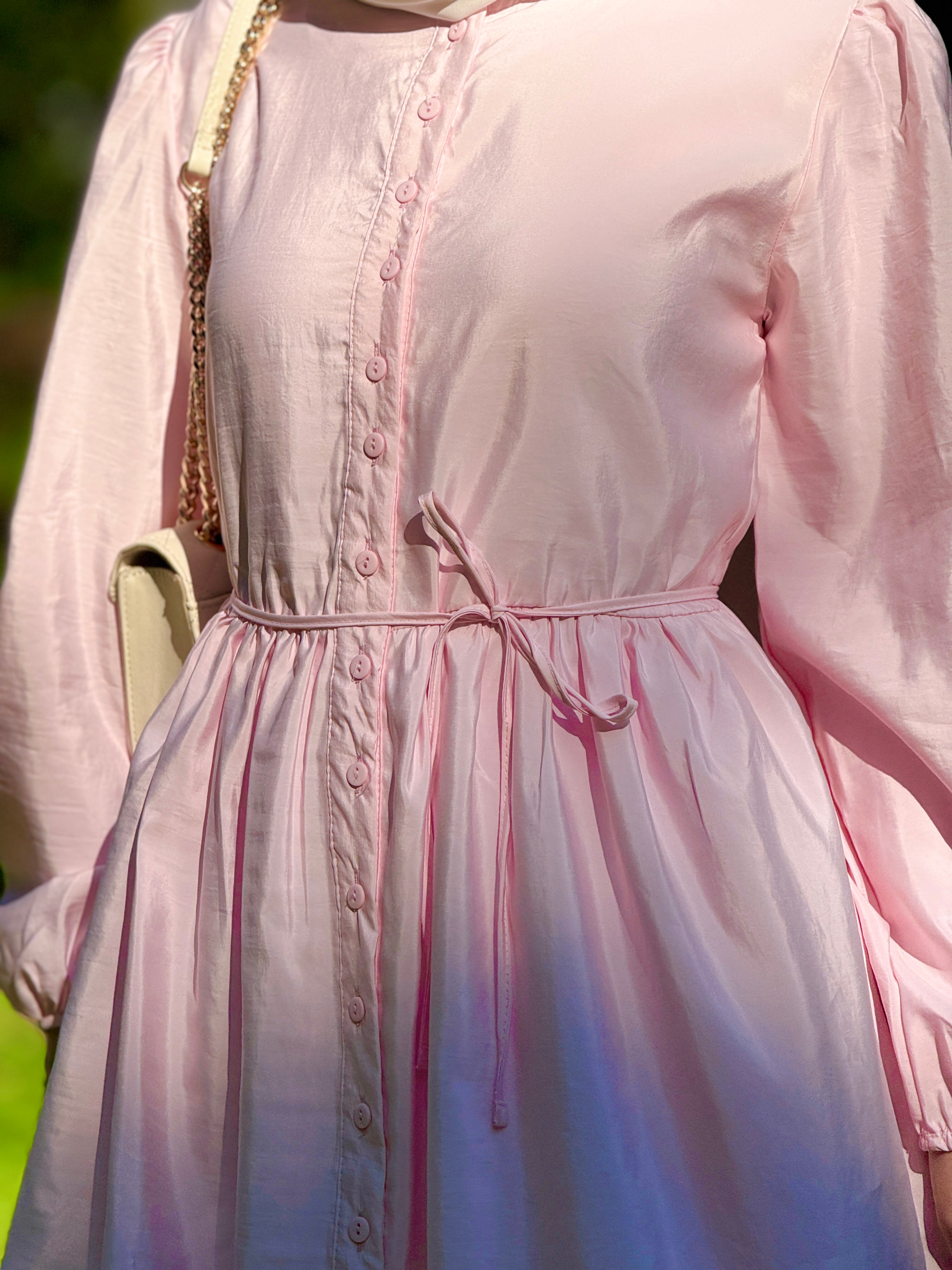 A close-up of a person wearing a light pink long-sleeve dress with a button front, gathered waist, and drawstring tie. A cream shoulder bag with a gold chain is partially visible.