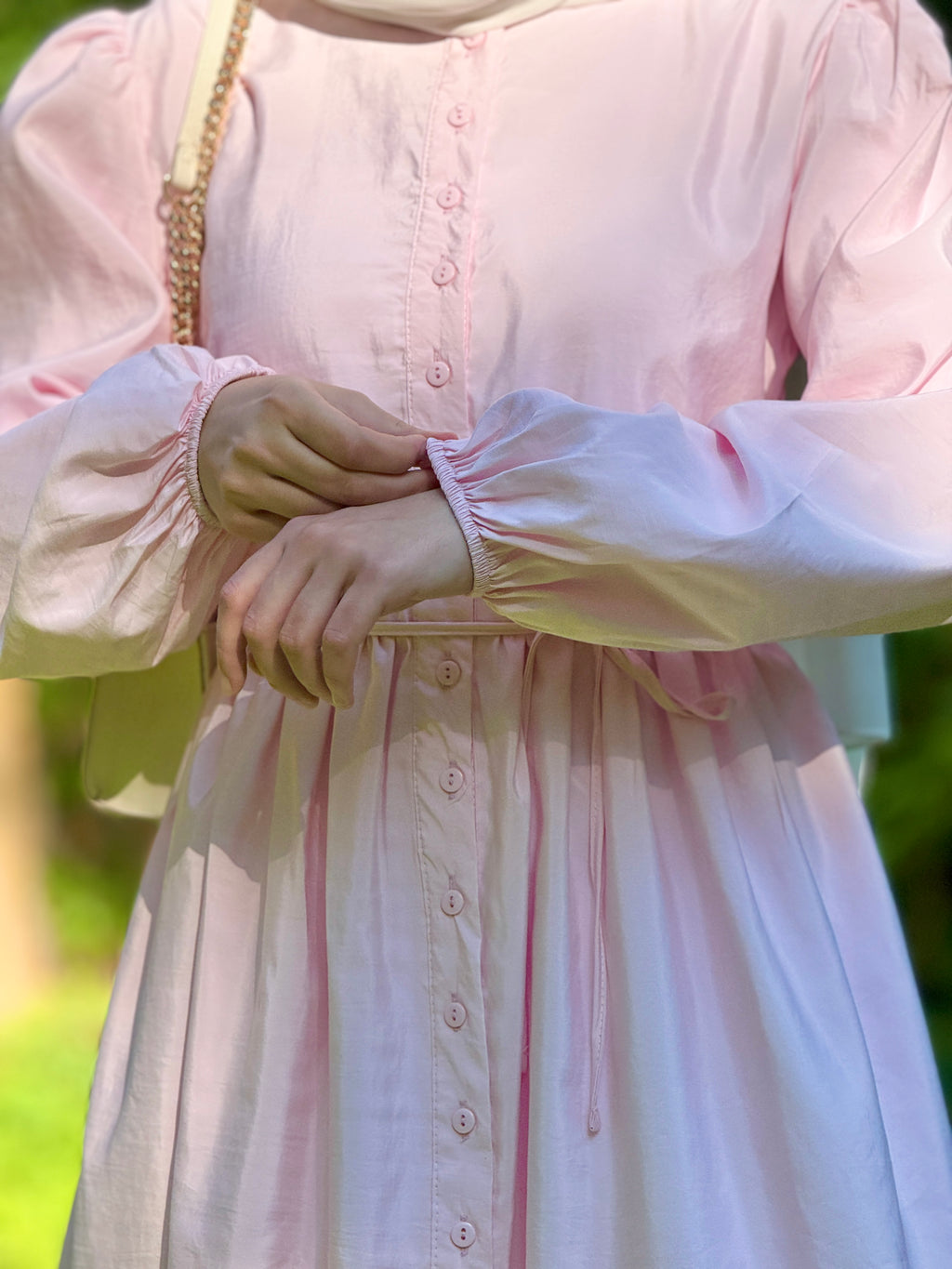 A close-up of a person wearing a light pink long-sleeve button-front dress with gathered cuffs and a drawstring waist, photographed outdoors with blurred greenery in the background.