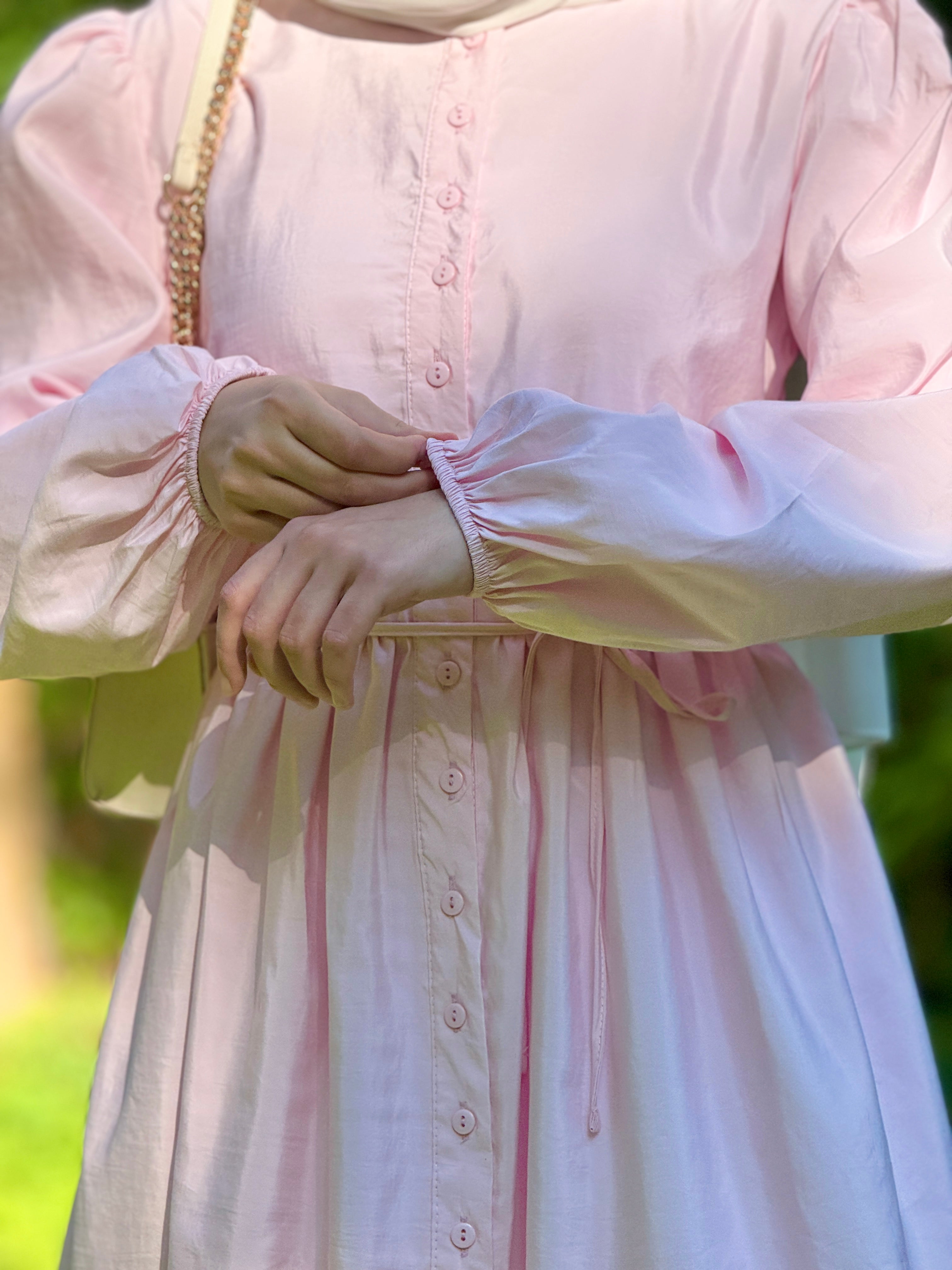 A close-up of a person wearing a light pink long-sleeve button-front dress with gathered cuffs and a drawstring waist, photographed outdoors with blurred greenery in the background.