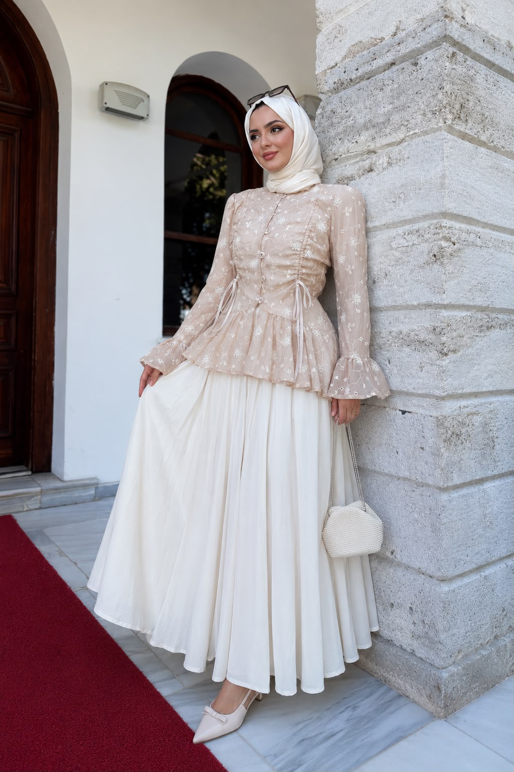 A woman wearing a beige and white modest outfit with a matching hijab, standing outdoors against a stone pillar next to a red carpet.