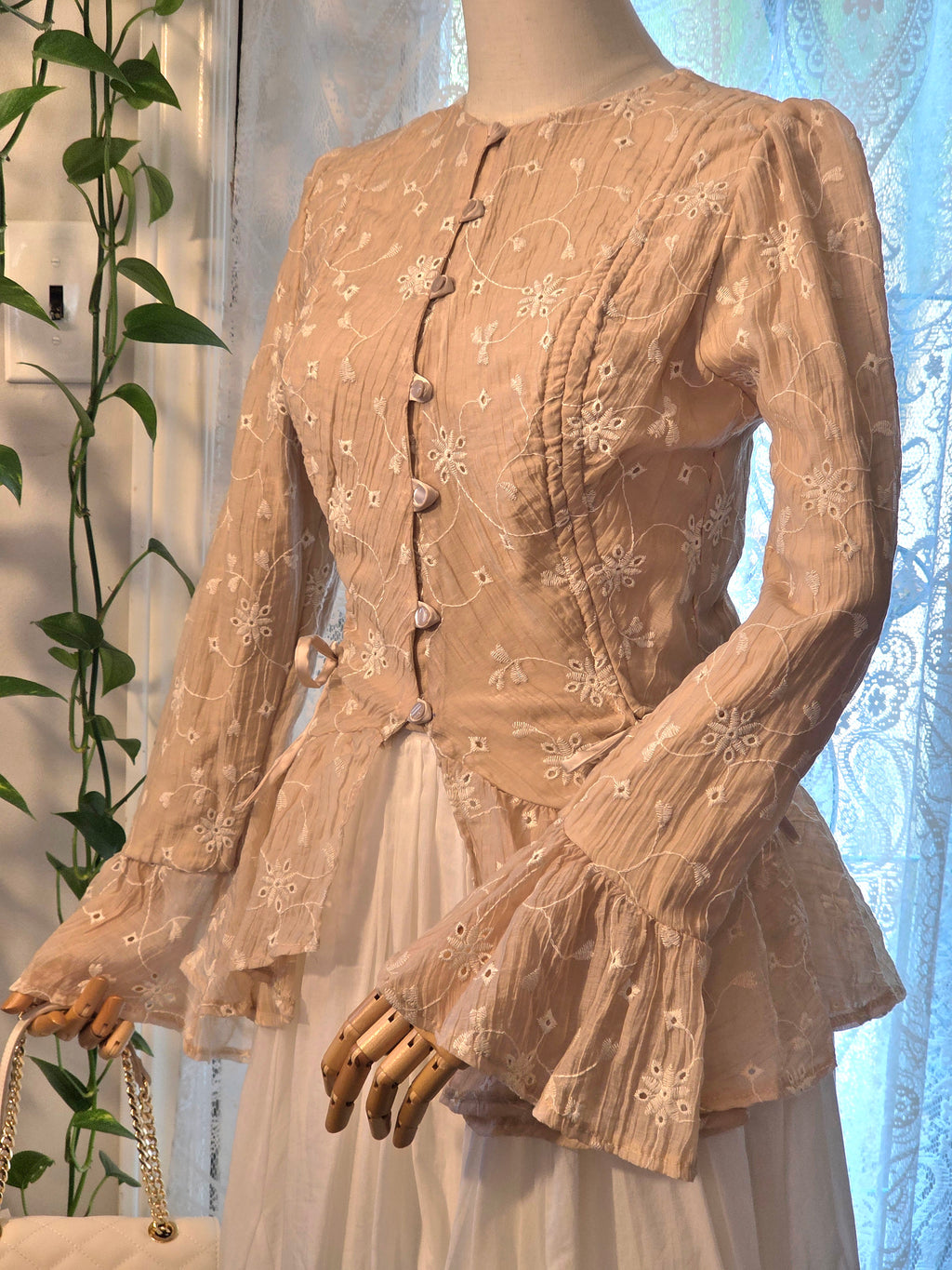 A beige long-sleeve blouse with white floral embroidery and decorative buttons, displayed on a mannequin with a white skirt. The blouse features a round neckline, fitted bodice, and flared ruffled sleeves.
