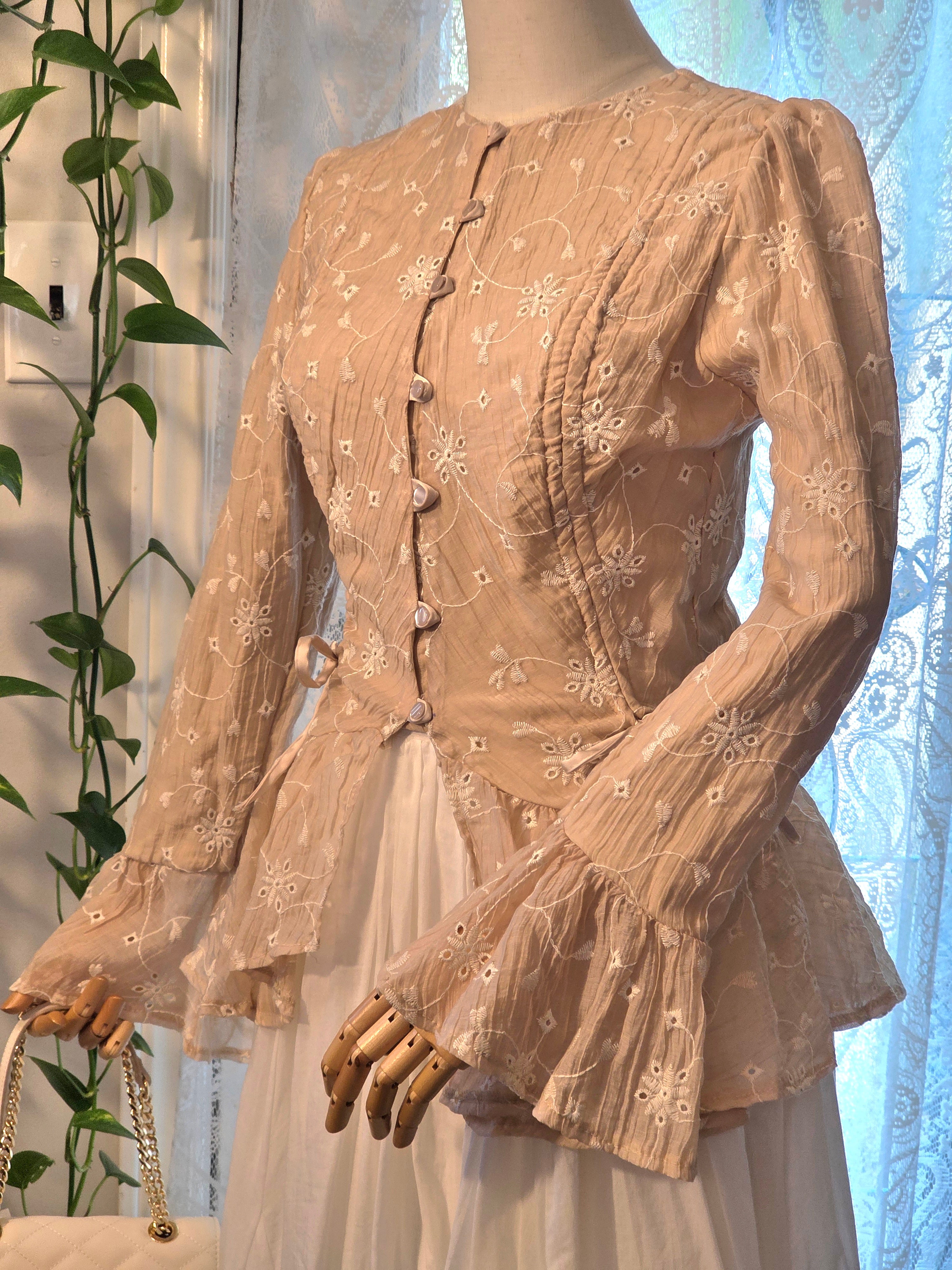 A beige long-sleeve blouse with white floral embroidery and decorative buttons, displayed on a mannequin with a white skirt. The blouse features a round neckline, fitted bodice, and flared ruffled sleeves.
