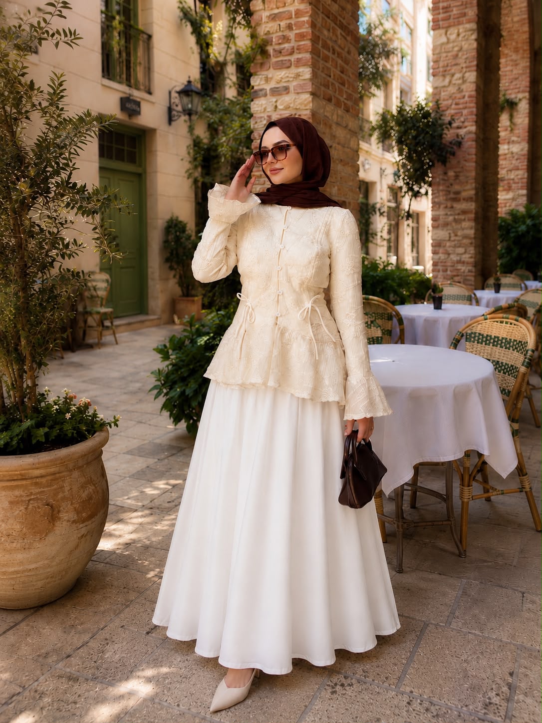 A woman wearing a cream lace long-sleeve peplum top and a white maxi skirt, styled with a dark brown hijab, sunglasses, and a small dark brown handbag, standing in an outdoor courtyard setting.