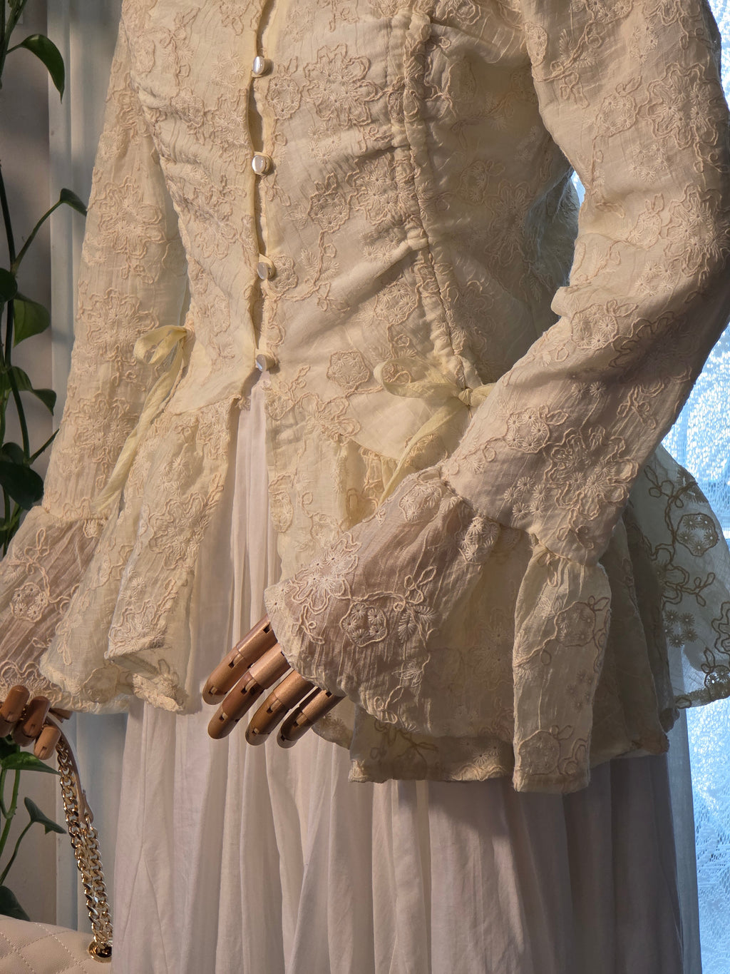 An off-white Victorian-style lace corset top with long bell sleeves and a buttoned front, displayed on a mannequin with a white skirt underneath.