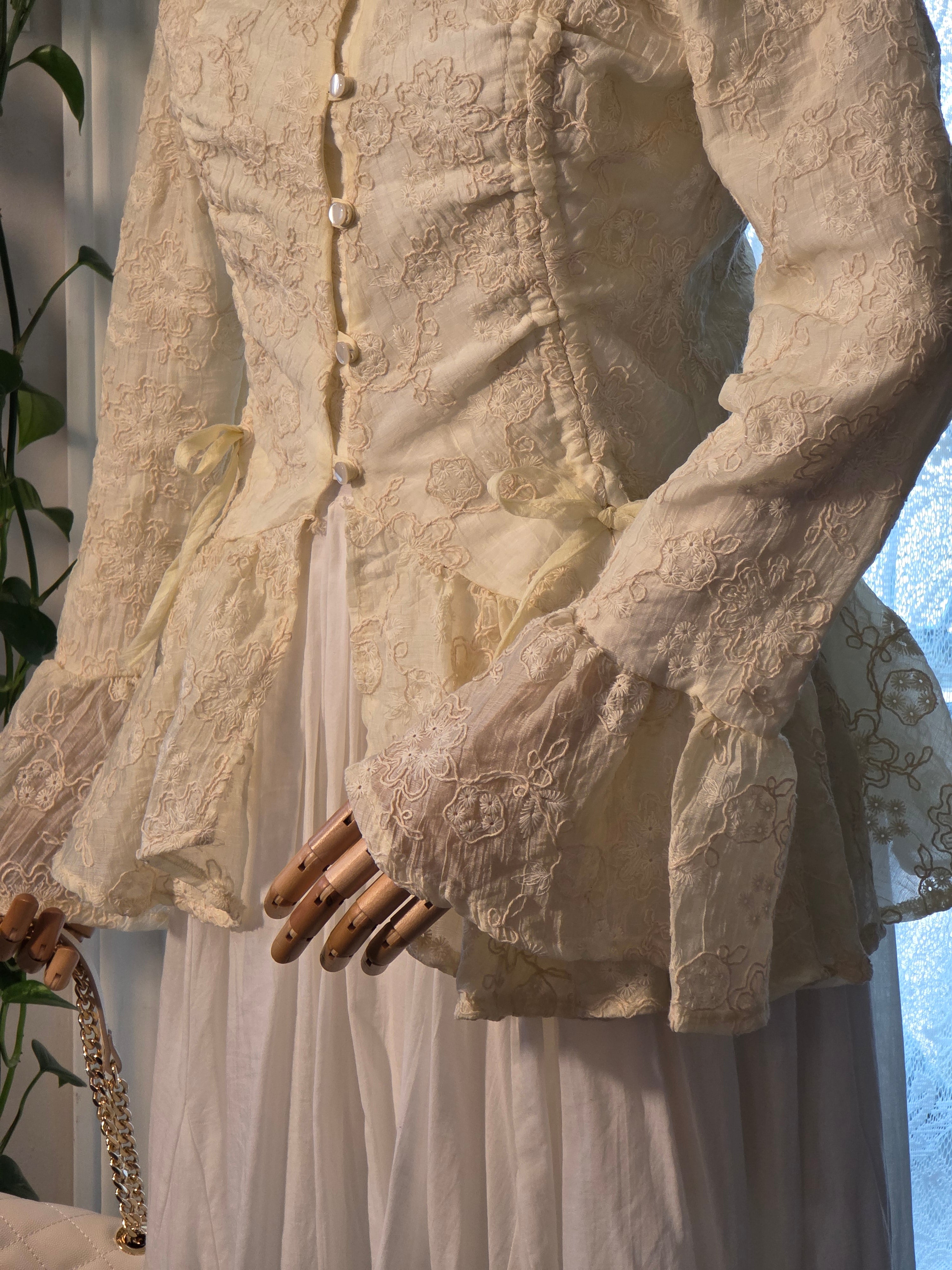 An off-white Victorian-style lace corset top with long bell sleeves and a buttoned front, displayed on a mannequin with a white skirt underneath.