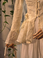 A close-up of a cream-colored lace and satin dress displayed on a mannequin, featuring long ruffled sleeves, a lace bodice with decorative buttons, and a flowing skirt. A wooden mannequin hand is visible, along with a gold chain handbag and green trailing vines against a neutral background.
