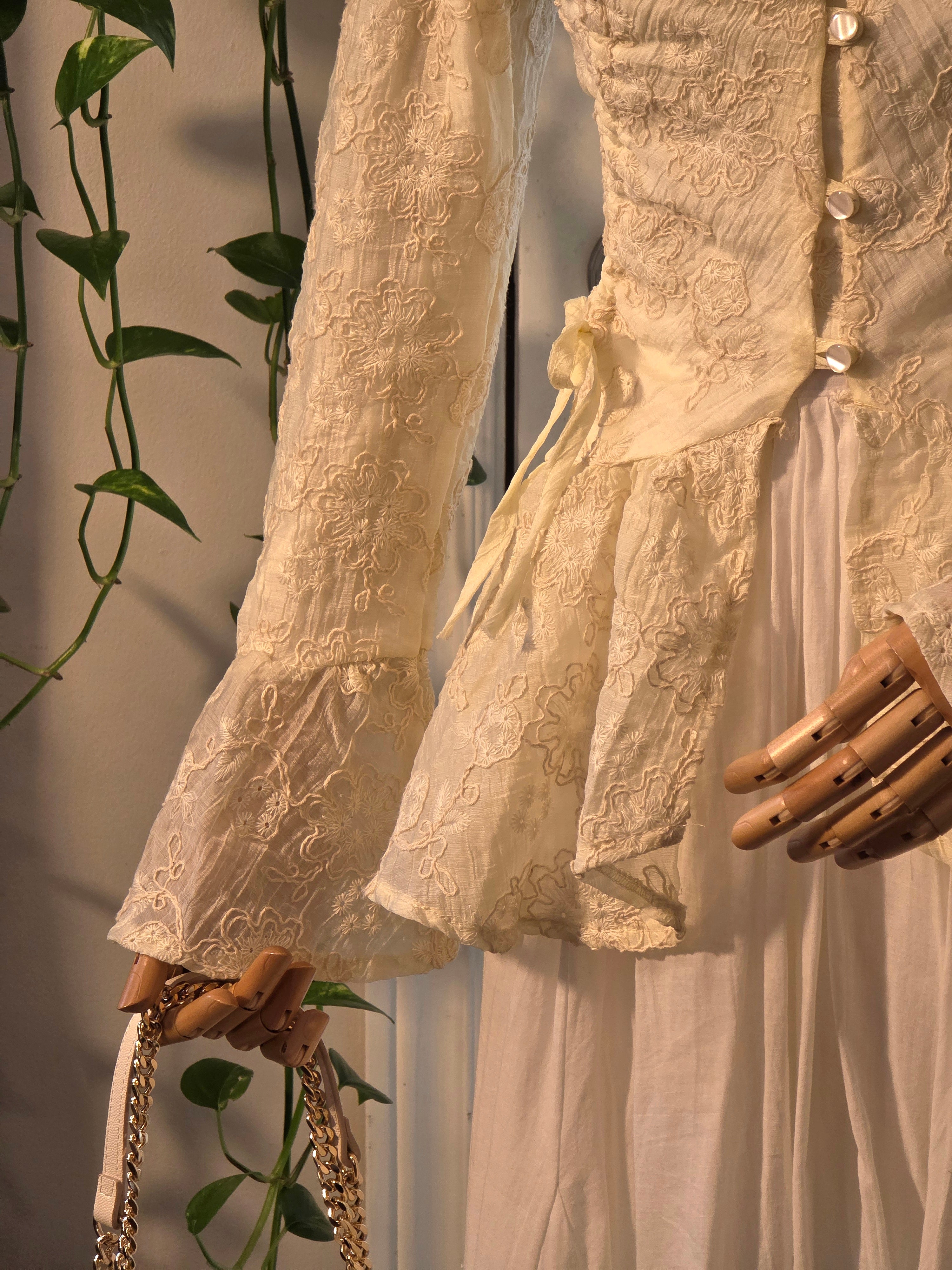 A close-up of a cream-colored lace and satin dress displayed on a mannequin, featuring long ruffled sleeves, a lace bodice with decorative buttons, and a flowing skirt. A wooden mannequin hand is visible, along with a gold chain handbag and green trailing vines against a neutral background.