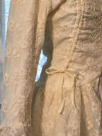 Close-up of a cream-colored lace dress with floral embroidery, a smocked bodice, and a matching waist tie, shown on a mannequin or hanger.