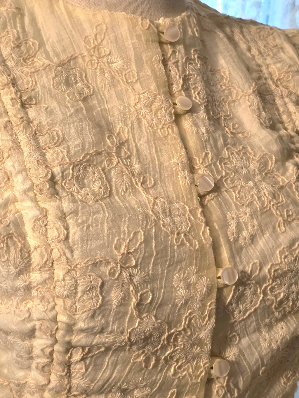 Close-up of a cream-colored textured fabric garment with floral embroidery and a buttoned front closure, displayed on a mannequin torso.