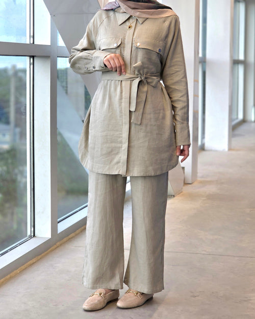 A woman models a matching beige linen two-piece set consisting of a long-sleeve button-up shirt with a tie waist belt and matching wide-leg trousers, paired with a beige hijab and beige loafers, standing in a modern indoor corridor with large windows.