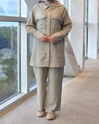 A person wearing a light beige linen two-piece outfit consisting of a long-sleeve button-up shirt with chest pockets and matching wide-leg trousers, standing indoors near a large window.