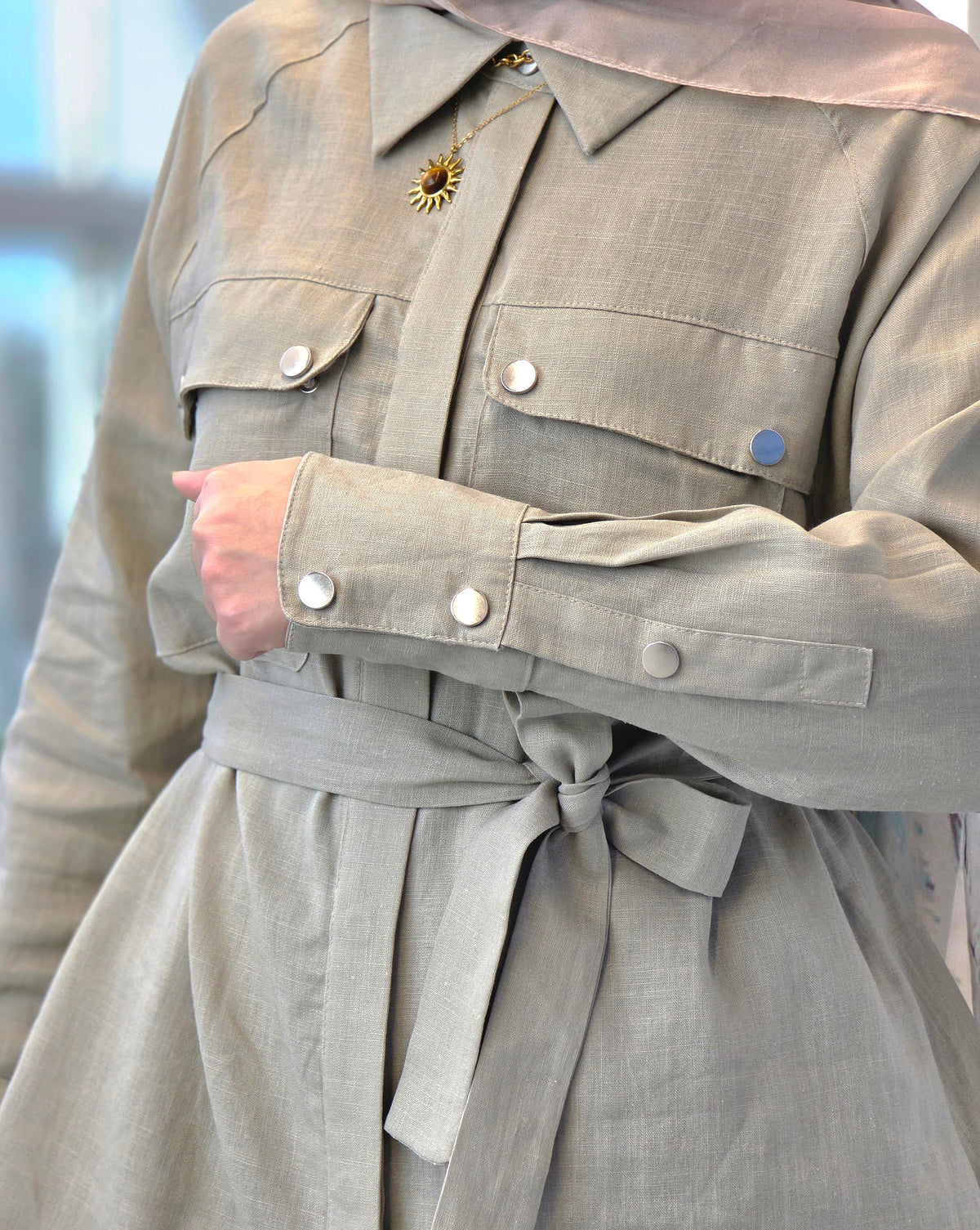 A close-up of a person wearing a beige linen shirt dress with a matching tie belt, featuring silver buttons and a gold sun pendant.