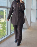 A person wearing a dark brown long-sleeve belted jacket and matching wide-leg trousers, standing indoors near large windows with a view of greenery outside.