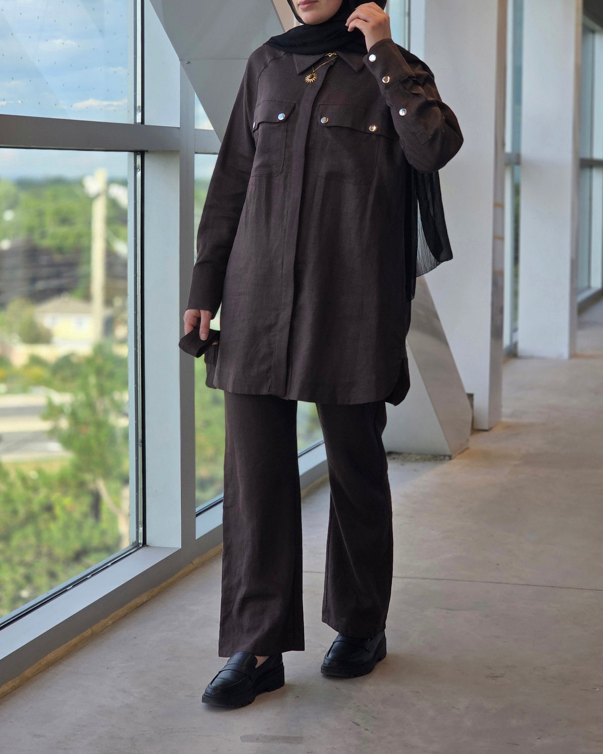 A woman wearing a dark brown long-sleeve button-up jacket, matching wide-leg trousers, and a black hijab stands in a modern indoor corridor with large windows.