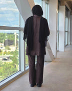 A person viewed from behind wearing a dark brown linen-like two-piece outfit with wide-leg pants and a sheer black draped shawl, standing in a modern indoor walkway next to large windows.
