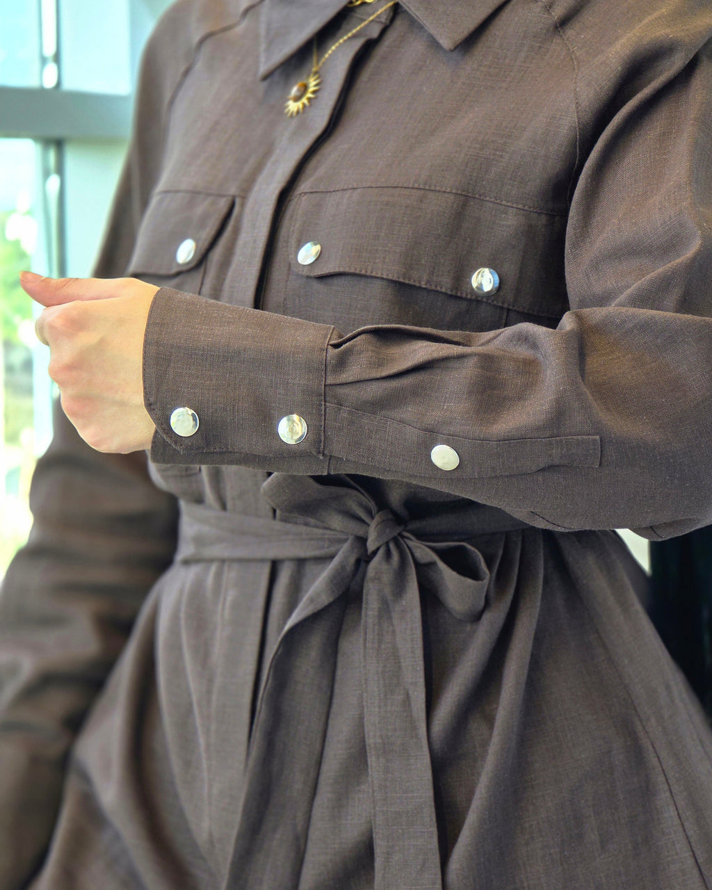 A close-up of a person wearing a dark brown long-sleeve shirt dress with silver snap buttons and a matching waist tie, paired with a black hijab.
