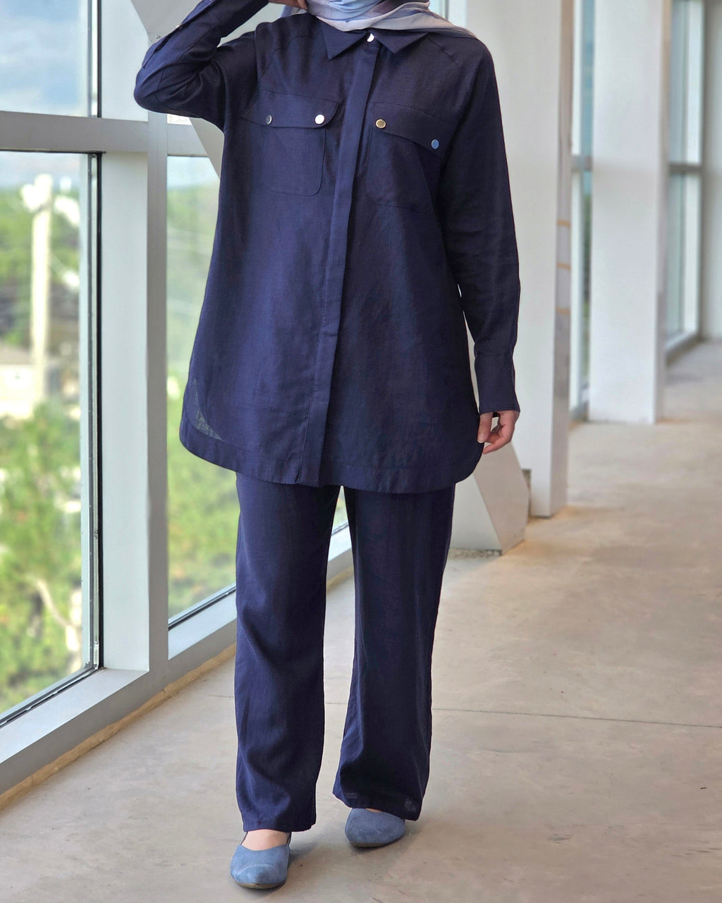 A person wearing a dark navy blue two-piece outfit consisting of a long-sleeve button-up shirt and matching wide-leg pants, standing in a modern indoor space with large windows.