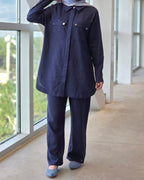 A person wearing a dark navy blue two-piece outfit consisting of a long-sleeve button-up shirt and matching wide-leg pants, standing in a modern indoor space with large windows.