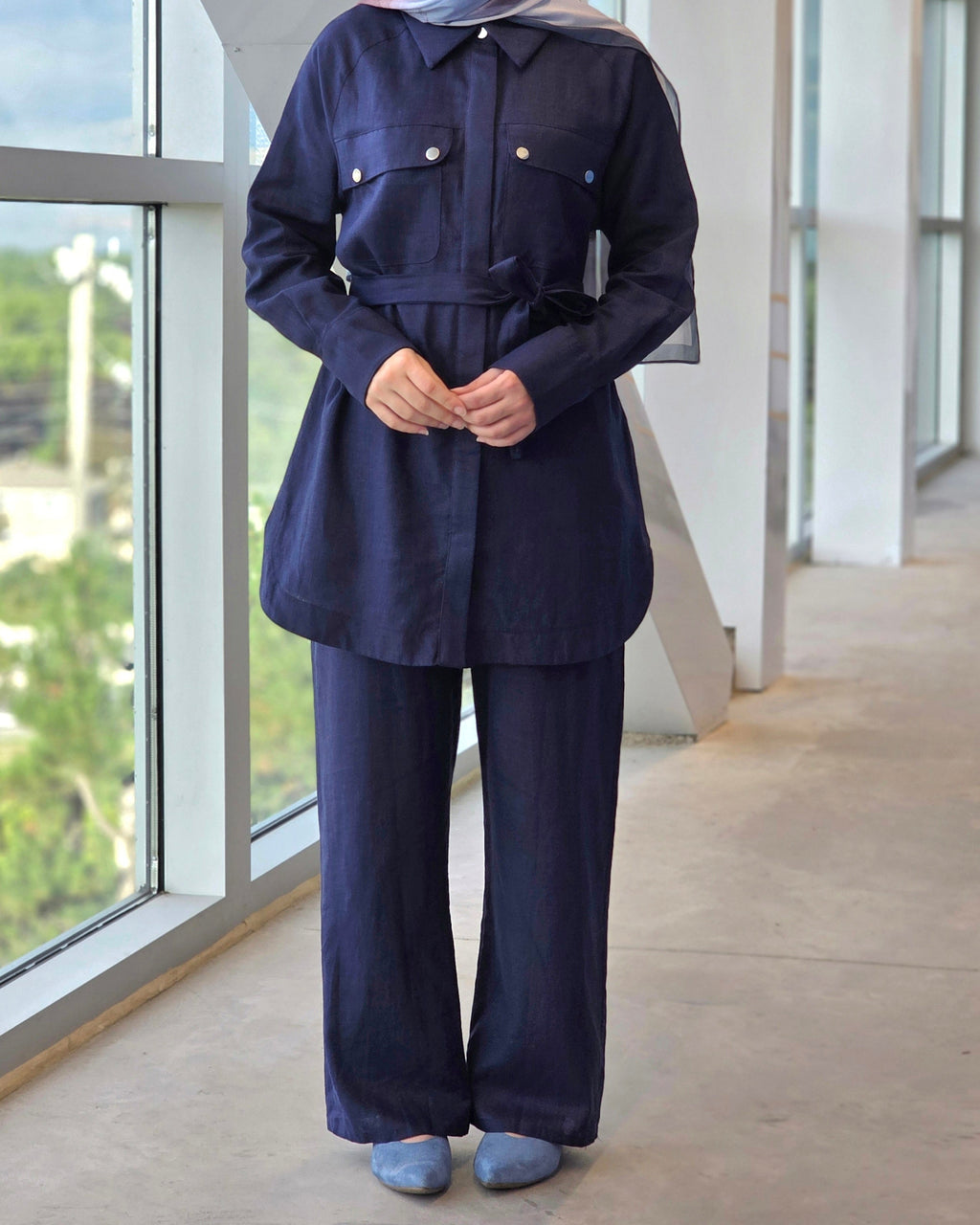 A person wearing a dark navy blue matching set consisting of a long-sleeve collared button-front top with a self-tie waist belt and matching wide-leg pants, paired with a patterned hijab and blue slip-on shoes, standing in a modern indoor corridor with large windows.