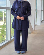 A person wearing a dark navy blue matching set consisting of a long-sleeve collared button-front top with a self-tie waist belt and matching wide-leg pants, paired with a patterned hijab and blue slip-on shoes, standing in a modern indoor corridor with large windows.