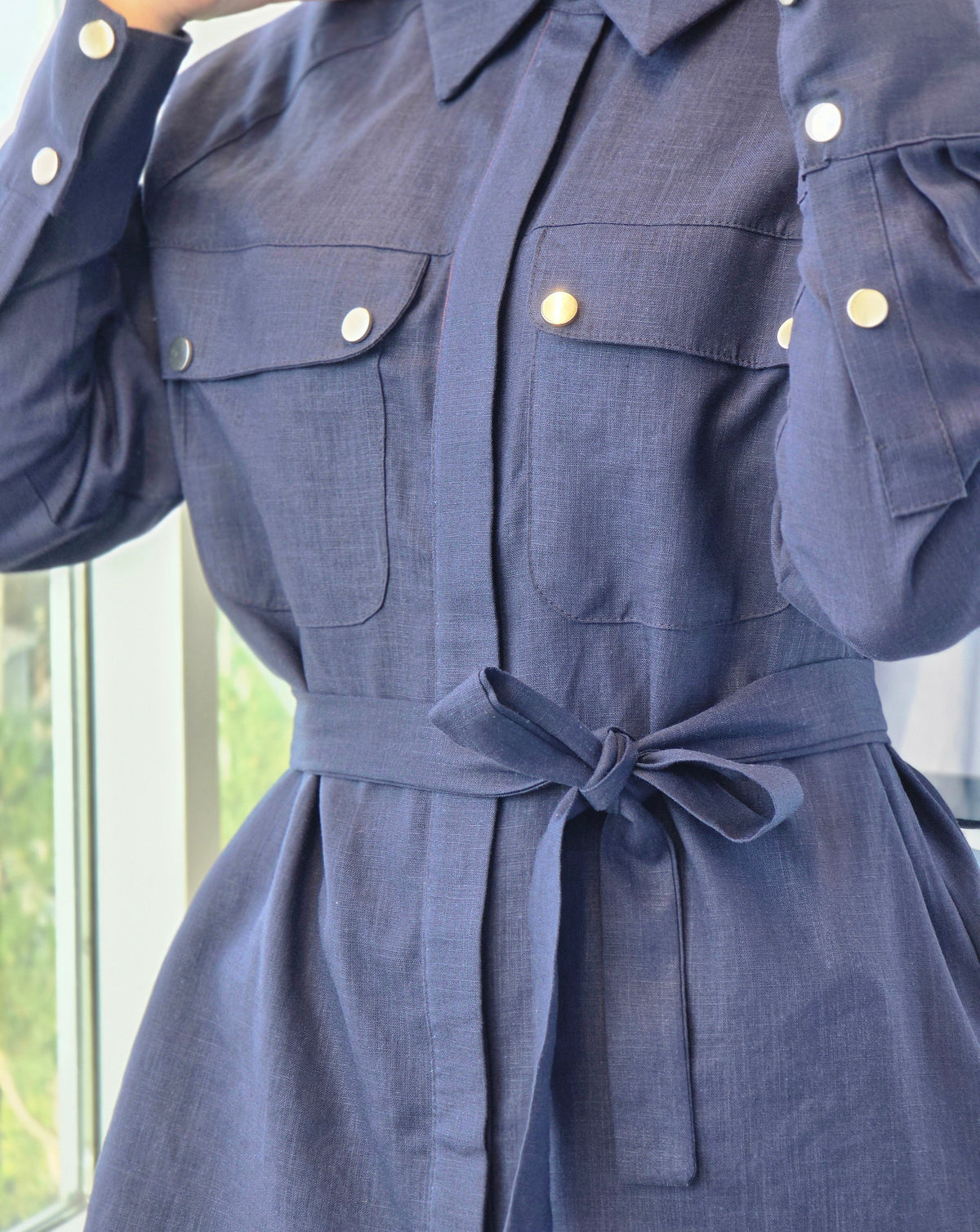 A close-up of a navy blue shirt dress with gold-tone snap buttons and a matching fabric tie belt at the waist, shown on a person indoors near a window.