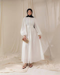 Woman wearing a white dress with long sleeves against a white curtain background