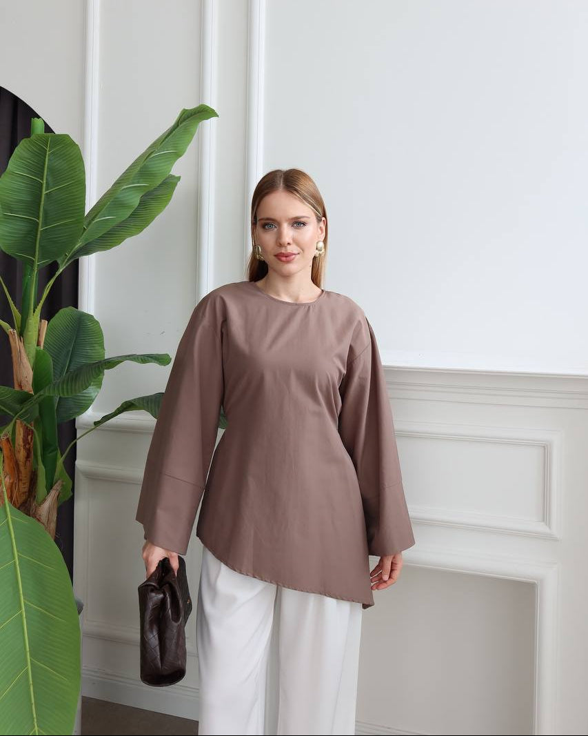 A woman modeling a taupe-brown long-sleeve top with white pants, standing in a bright room with a white paneled wall and a large green plant on the left.
