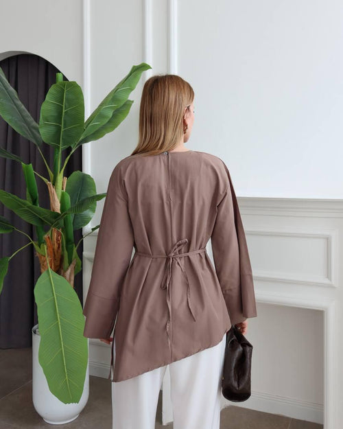 A woman viewed from behind wearing a taupe-brown long-sleeve wrap-style top with a drawstring waist, paired with white pants, standing in a bright room next to a potted green plant.
