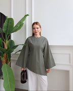 A woman wearing a muted sage green long-sleeve top with wide bell sleeves and white pants, holding a small dark brown handbag. She is standing in a bright room with white walls and molding next to a large green potted plant.
