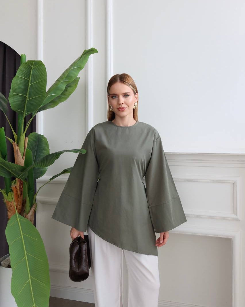 A woman wearing a muted sage green long-sleeve top with wide bell sleeves and white pants, holding a small dark brown handbag. She is standing in a bright room with white walls and molding next to a large green potted plant.