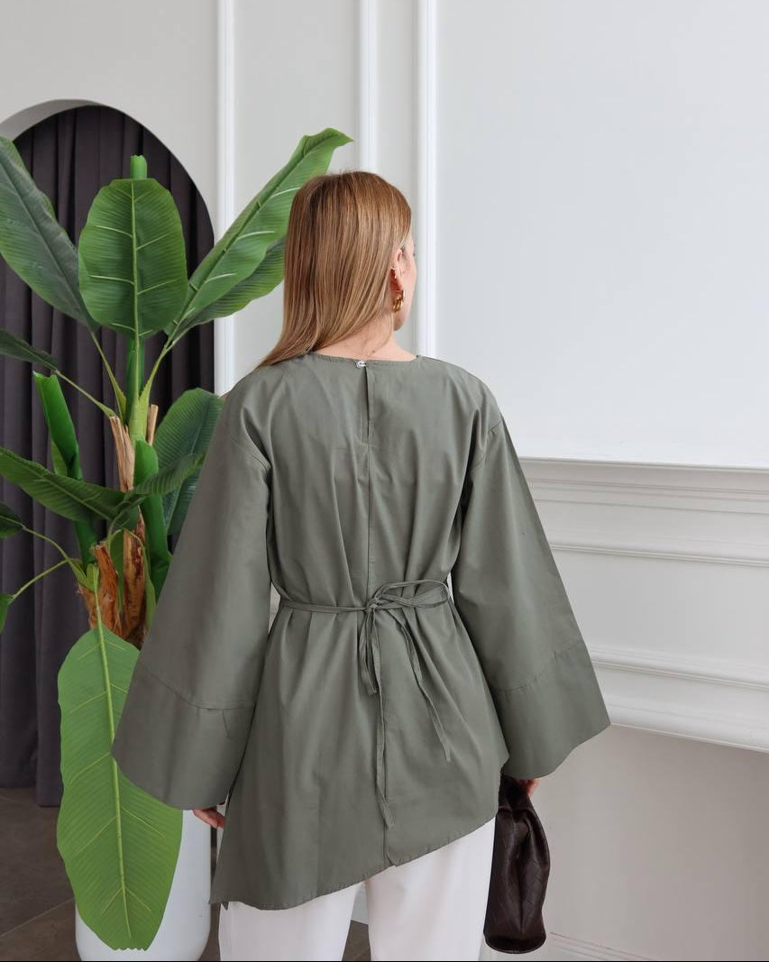 A woman viewed from behind wearing a loose, short-sleeved sage green top with a drawstring waist, paired with white pants, standing in a bright room next to a large potted plant.