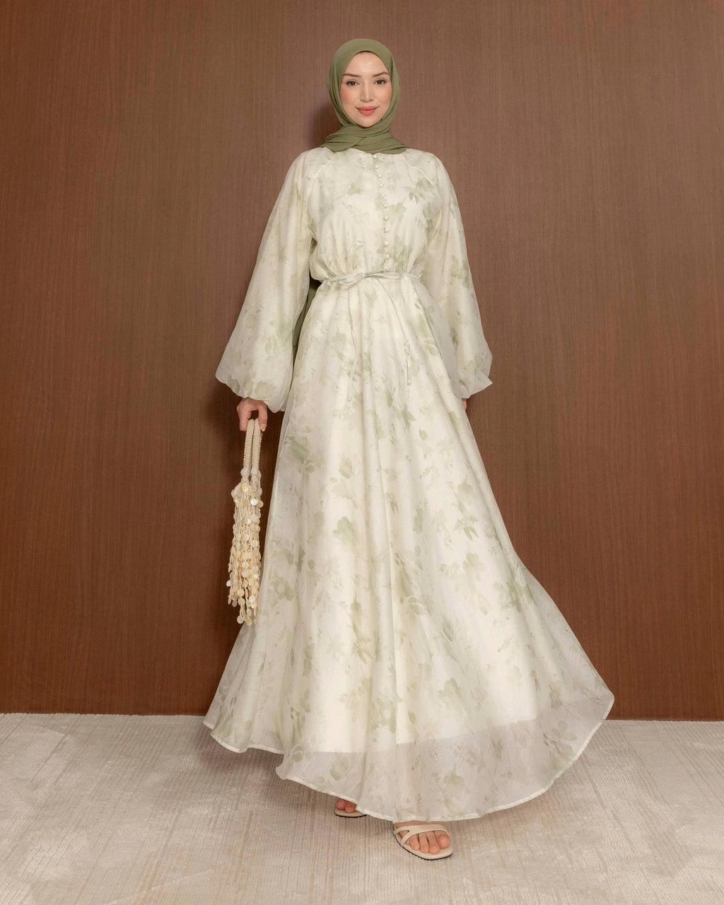 A woman wearing a long, flowing cream-colored maxi dress with pastel green flowers with a matching headscarf, holding a small woven bag, standing against a brown wooden wall.