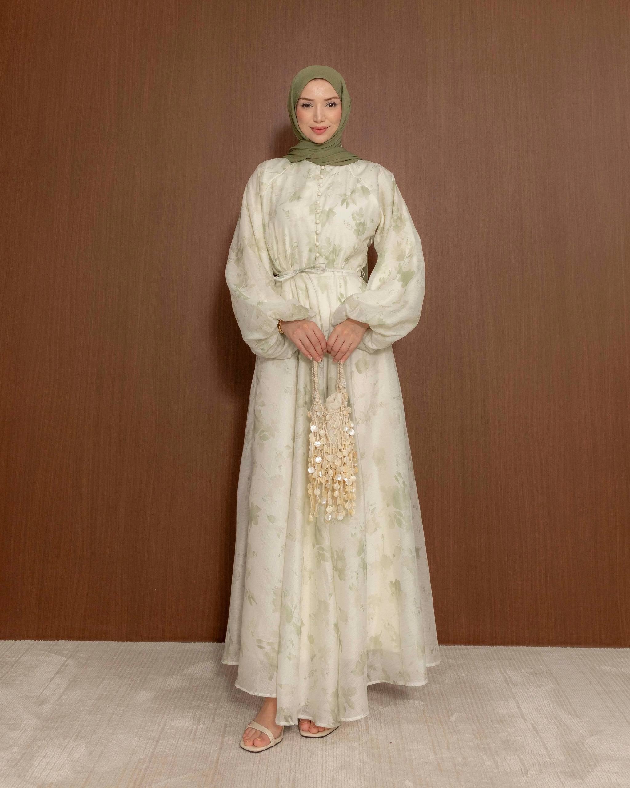 A woman wearing a long, flowing cream-colored modest dress with a light green floral pattern, a matching green hijab, and beige sandals, standing against a brown wood-paneled wall.