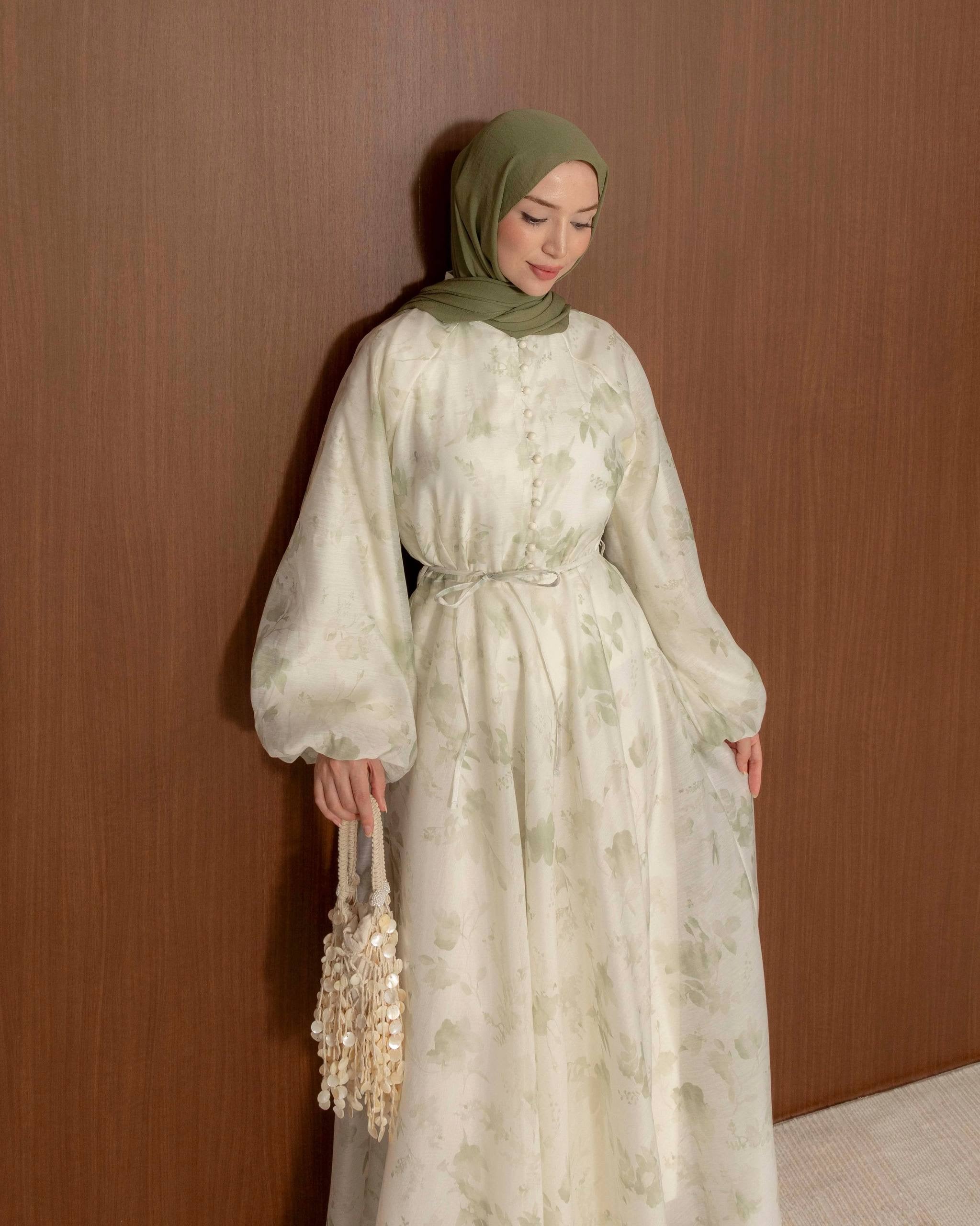 A woman wearing a long, flowing cream-colored dress with a light sage green floral pattern, a matching sage green hijab, and holding a beaded handbag, standing against a brown wooden wall.