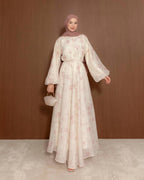 A woman wearing a long, flowing cream-colored modest dress with a light pink floral pattern, a matching pink hijab, and holding a small cream handbag, standing against a brown wooden wall.